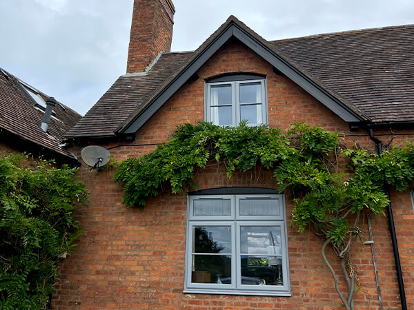 Two cottage windows. Both High Security, Extreme, Hazy Grey, UPVC double glazed Windows fitted into a wisteria clad gable end of a red brick house.